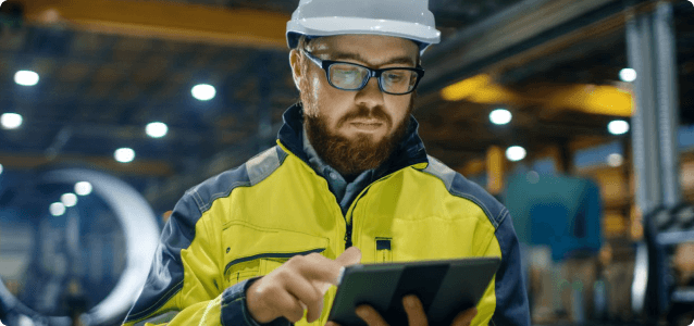 Man in hardhat, safety glasses, and hi-vis jacket interacts with tablet in a warehouse